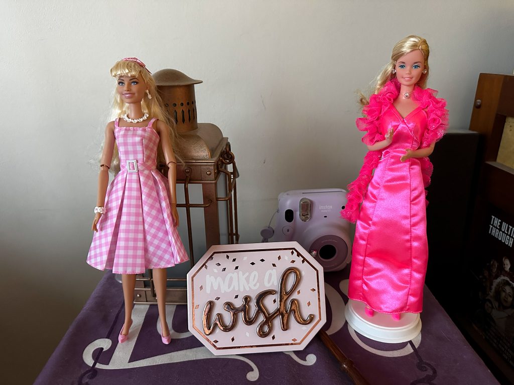Two Barbie dolls on a shelf with a sign between them.