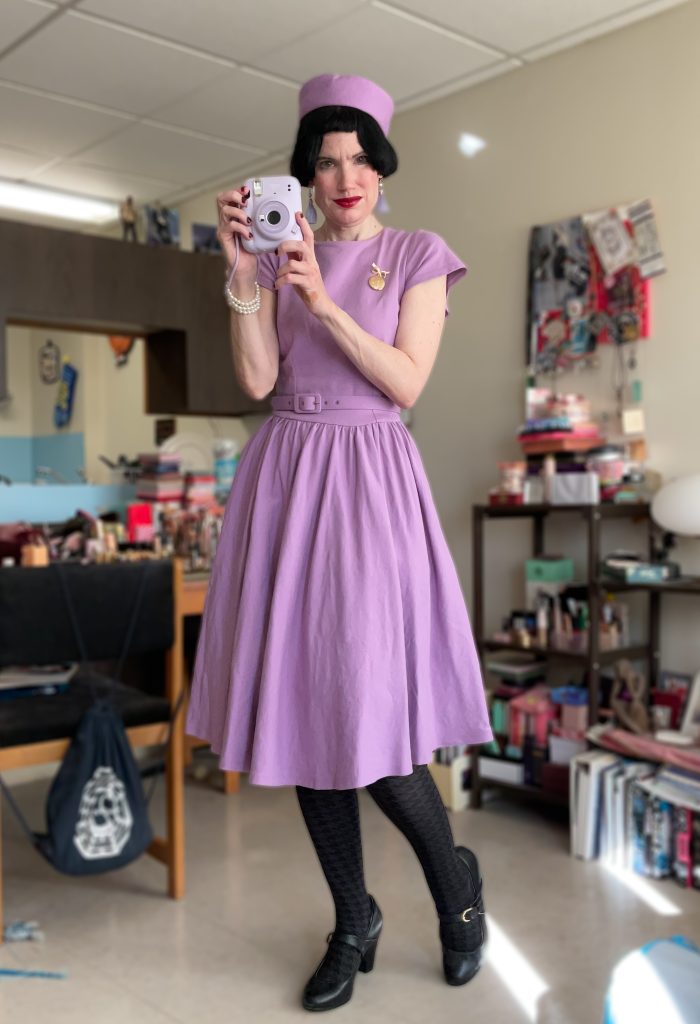 Me in lavender dress and pillbox hat holding a camera